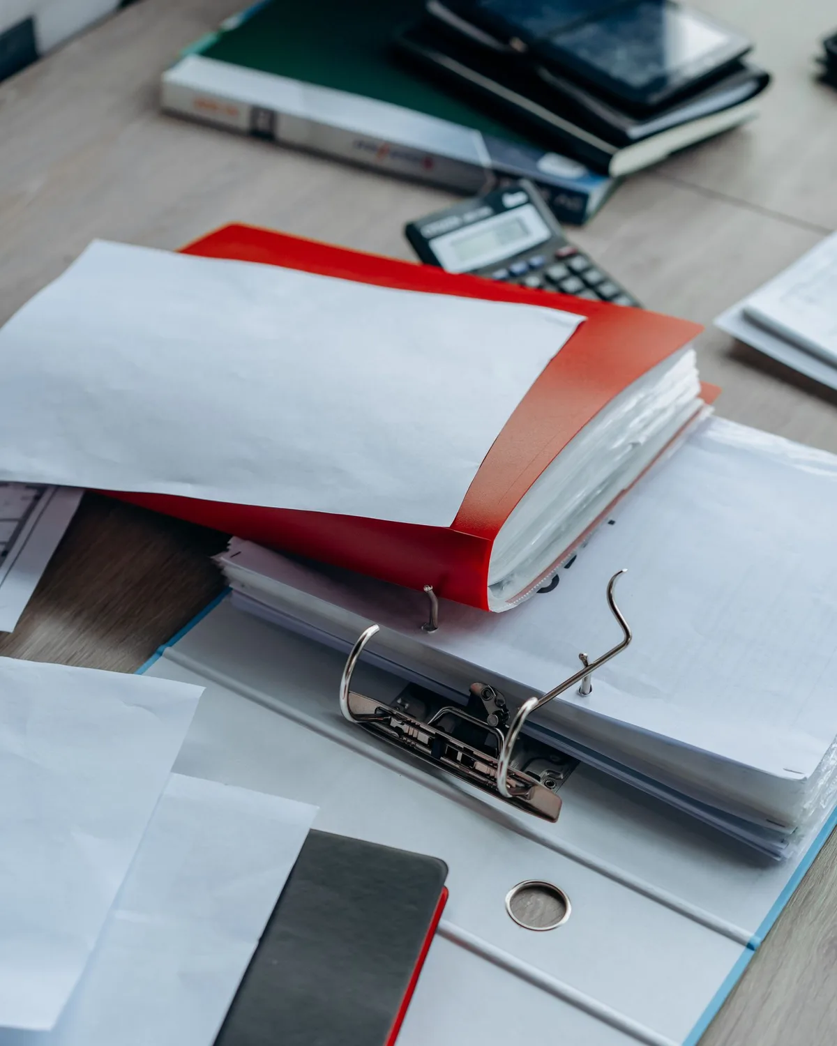 書類がはさまれた赤と白のリングファイル、電卓、黒い手帳が置かれた机