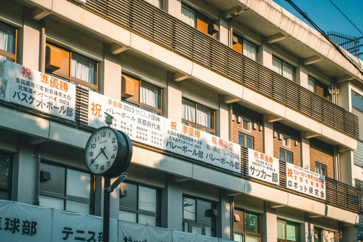 部活動の垂れ幕が並ぶ高校の校舎と、校門の前に立つ丸い時計